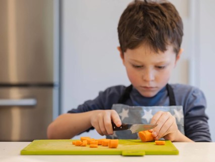 A focused young child in a blue apron chops a carrot on a green cutting board. The image captures