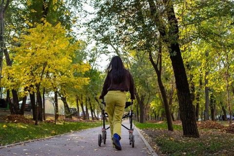 A woman with cerebral palsy walks with a walker on a scenic park path surrounded by vibrant autumn