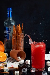 Splashing long drink with vodka and oranges, surrounded by cards, dice and pretzel sticks, studio