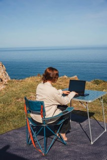 A woman enjoys remote work while sitting on a chair near the ocean, surrounded by stunning views.