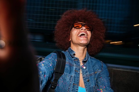 A joyful African descent woman with afro hair, wearing orange sunglasses, laughs heartily in an