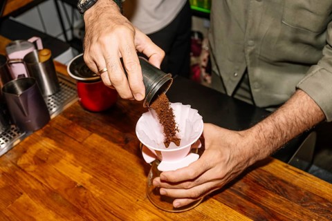 A barista pours freshly ground coffee into a paper filter placed in a pour-over coffee maker. The
