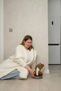 A woman wearing a cozy sweater sits on the floor, feeding her small dog from a bowl. The atmosphere