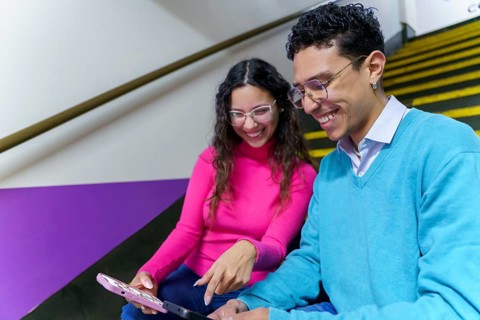 A cheerful woman and a man sitting on a staircase, smiling while interacting with a tablet. They