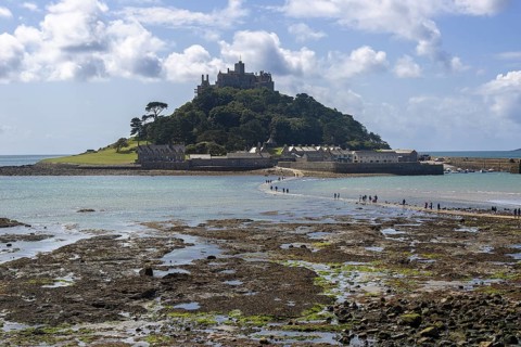 St Michael's Mount on an island, stone beach with people in the foreground, blue sky with clouds,