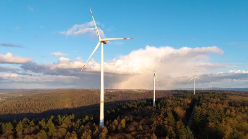 Tall wind turbines in a wooded landscape stretching into the distance under a clear sky, wind