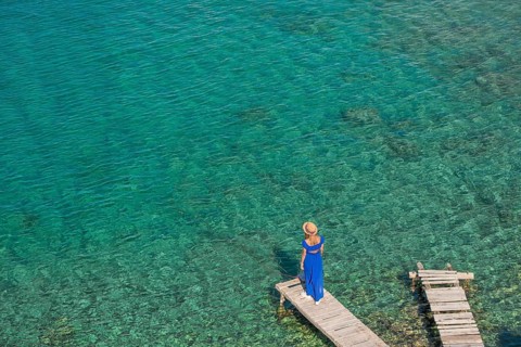 A woman in a flowing blue dress and hat stands on a wooden dock of Mandrakia, Milos island, Greece.