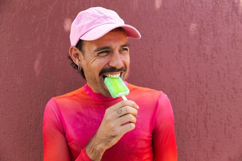 A young Spanish LGTBIQ+ man with a mustache showcases lively urban fashion as he enjoys a popsicle