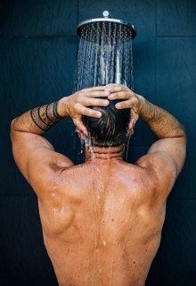 A man with tattoos enjoys a refreshing shower, standing with his back to the camera. Water cascades