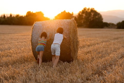 Back view of two unrecognizable children, a boy and a girl, play by pushing a large hay bale in a