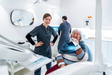 Happy elderly patient sitting in dentist chair while doctor and nurse smiling in modern bright