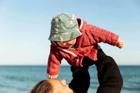 A mother joyfully lifts her child at the beach in Samalus, Barcelona. The toddler wears a cozy red