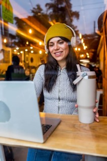 Woman wearing a beanie and headphones, holding a reusable tumbler while working on a laptop at a
