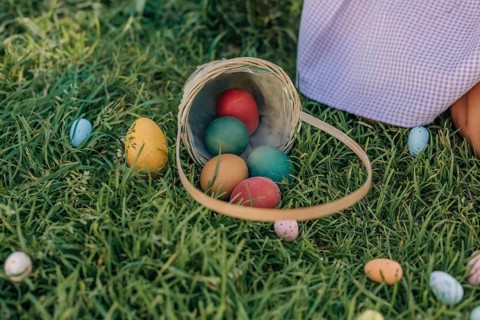 A wicker basket overflows with colorful Easter eggs on lush green grass. Pastel eggs are scattered,