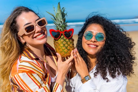 Two smiling young women friends holding a pineapple wearing red heart shaped sunglasses, enjoying