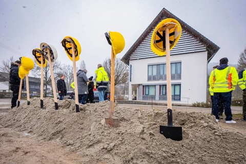 Yellow helmets on spades stuck in a pile of sand, with people and buildings in the background,