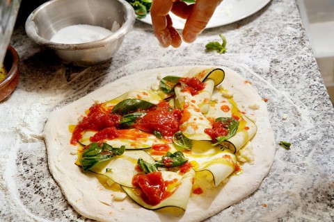 Cropped unrecognizable person hands adds toppings to a fresh pizza dough with zucchini slices,