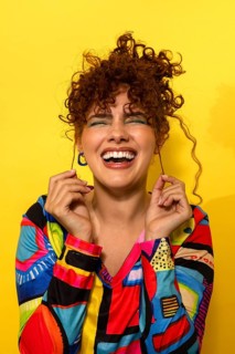 A joyful woman with curly hair laughs heartily while holding her hair. She wears a colorful