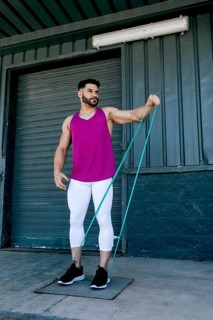 A fit man in a pink tank top and white leggings exercises with a resistance band outdoors. He
