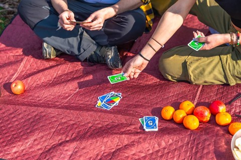 Hikers enjoying a lively card game while relaxing on a red quilted blanket, surrounded by fresh