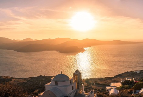 Sun sets behind Greek island, casting golden hues over the Aegean Sea. A white domed church is
