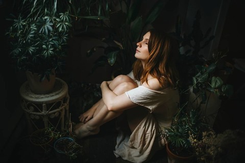 A serene scene of a woman sitting among lush indoor plants, bathed in soft light. The ambiance is