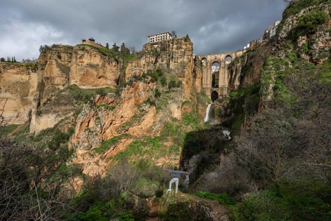 A traveler stands on a rock overlooking a waterfall and the massive arches of the Puente Nuevo