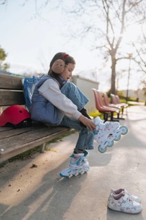 Side view of a young girl sits on a wooden bench in a sunlit park, putting on her roller skates,