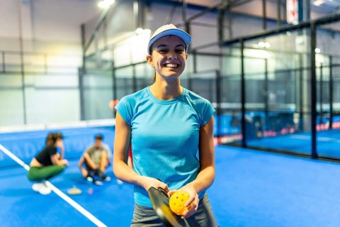 Woman smiling at camera while holding a pickleball paddle and ball on a blue indoor court,