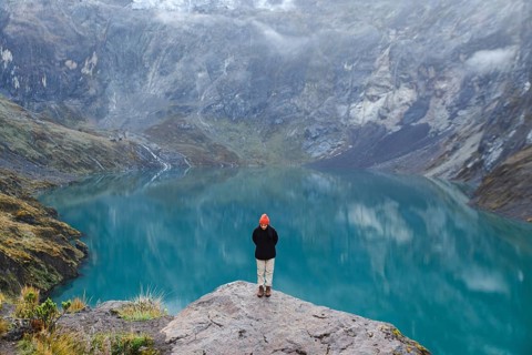 An Ecuadorian woman stands on a rocky ledge overlooking a serene turquoise lake at El Altar Volcano