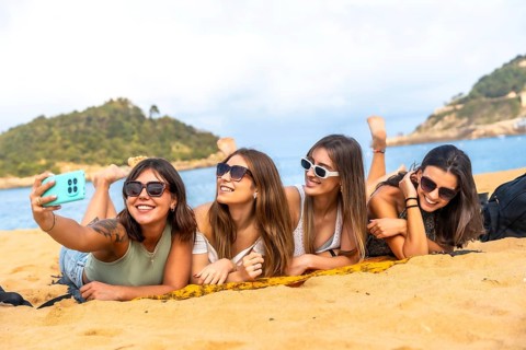 Four young women friends lying on a sandy beach and taking a selfie with a smartphone, enjoying