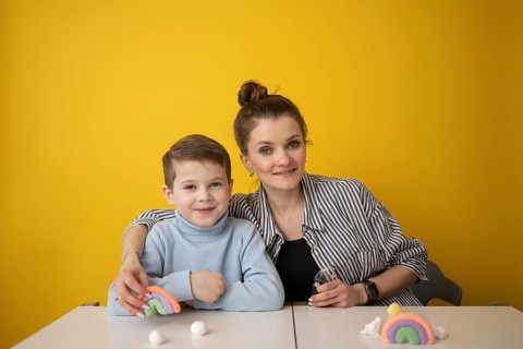 A child and a woman smile in front of a yellow wall, enjoying a creative lesson with colorful