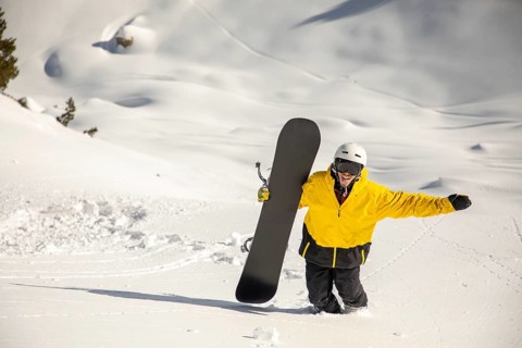 A snowboarder in a bright yellow jacket treks through powdery snow, holding a snowboard and ready
