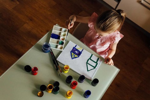A young girl sits at a table, focused on painting a colorful picture in a sketchbook. Surrounded by