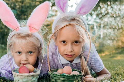 Two children wearing bunny ears lie on grass with baskets of colorful Easter eggs, enjoying a sunny