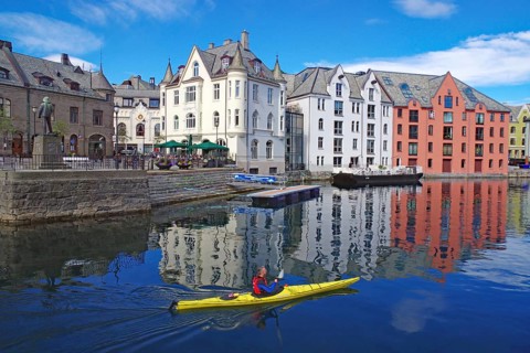 A kayaker paddles past colorful buildings that are reflected in the water in a harbor, Art Nouveau,