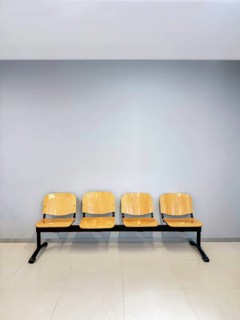 A row of four wooden chairs with metal frames placed against a light gray wall. The minimalist