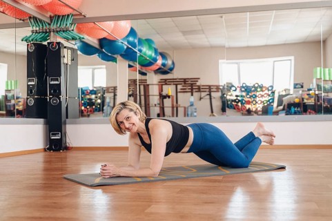 A smiling mature woman is performing a modified plank exercise on a yoga mat in a gym, smiling at