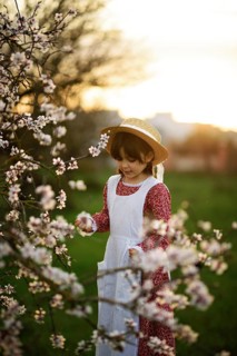 A young girl in a vintage dress and straw hat enjoys a serene moment in a blossoming almond tree at