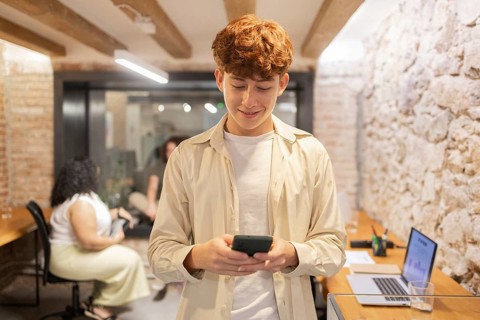 A young man with red hair is focused on his smartphone in a trendy coworking space, while others