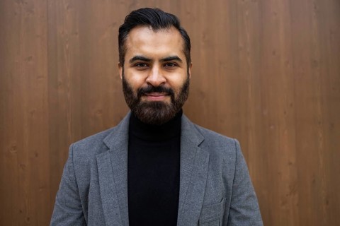 A well-dressed Indian man in business attire stands confidently against a wooden background in his