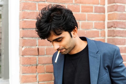 A indian man in a blue blazer stands against a brick wall, smoking. The casual pose and soft