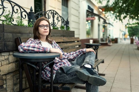 A teenage boy with long hair and glasses sits casually in an outdoor cafe setting, wearing a plaid