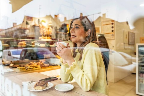 Young woman happily drinking coffee and looking away while sitting at a wooden table with a pastry,