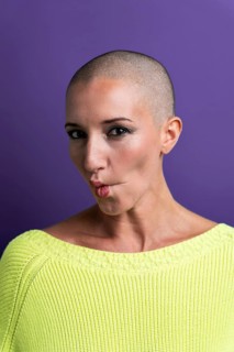 A middle-aged woman poses confidently with a shaved head, wearing a bright yellow sweater. Her