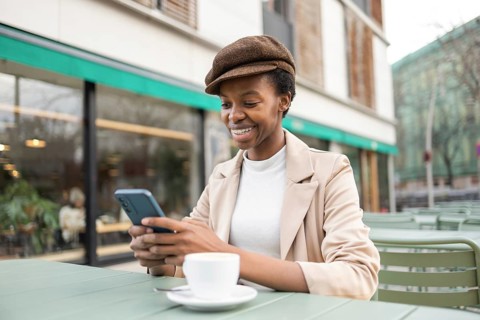 A black woman in an urban cafe setting, smiling while using her phone She wears a stylish hat and