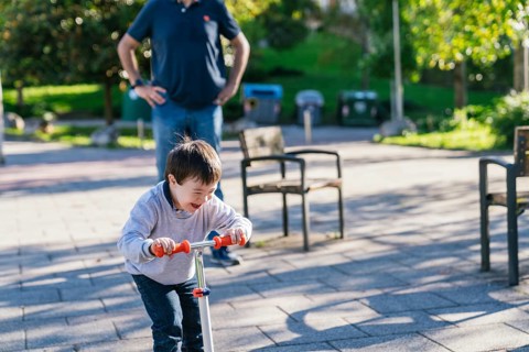 A cheerful child with Down syndrome rides a scooter through a sunlit park, laughing as his father