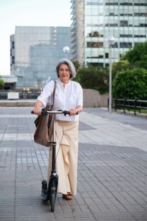 Mature woman confidently rides an electric scooter in a modern urban environment. She wears a white