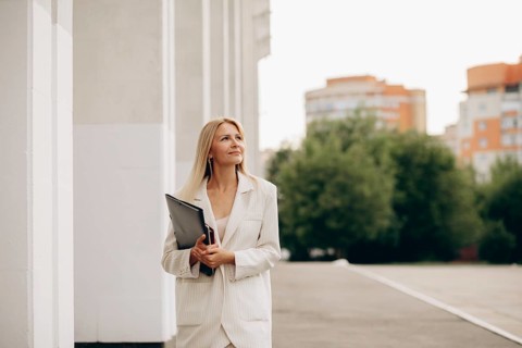 A businesswoman in a white suit holds a folder while walking on a city street. Urban buildings and