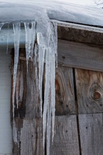 Long icicles hanging from snow and ice covered roof edge of old wood plank cladded building in